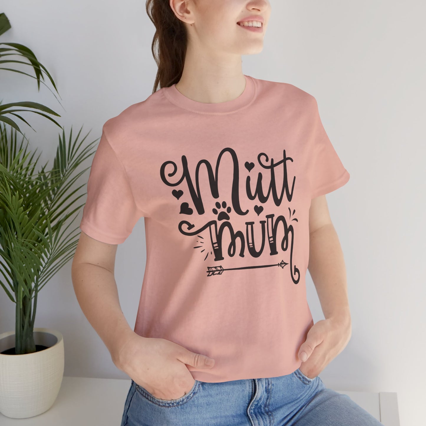 Person wearing a pink t-shirt with 'Mutt Mum' text, standing indoors next to a plant.