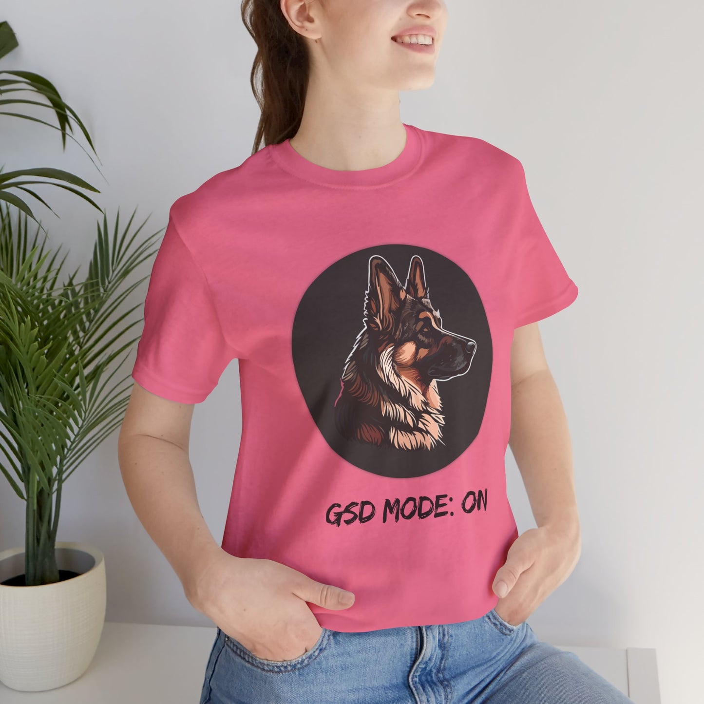 Person wearing a pink t-shirt with a dog graphic and text, standing indoors next to a plant.