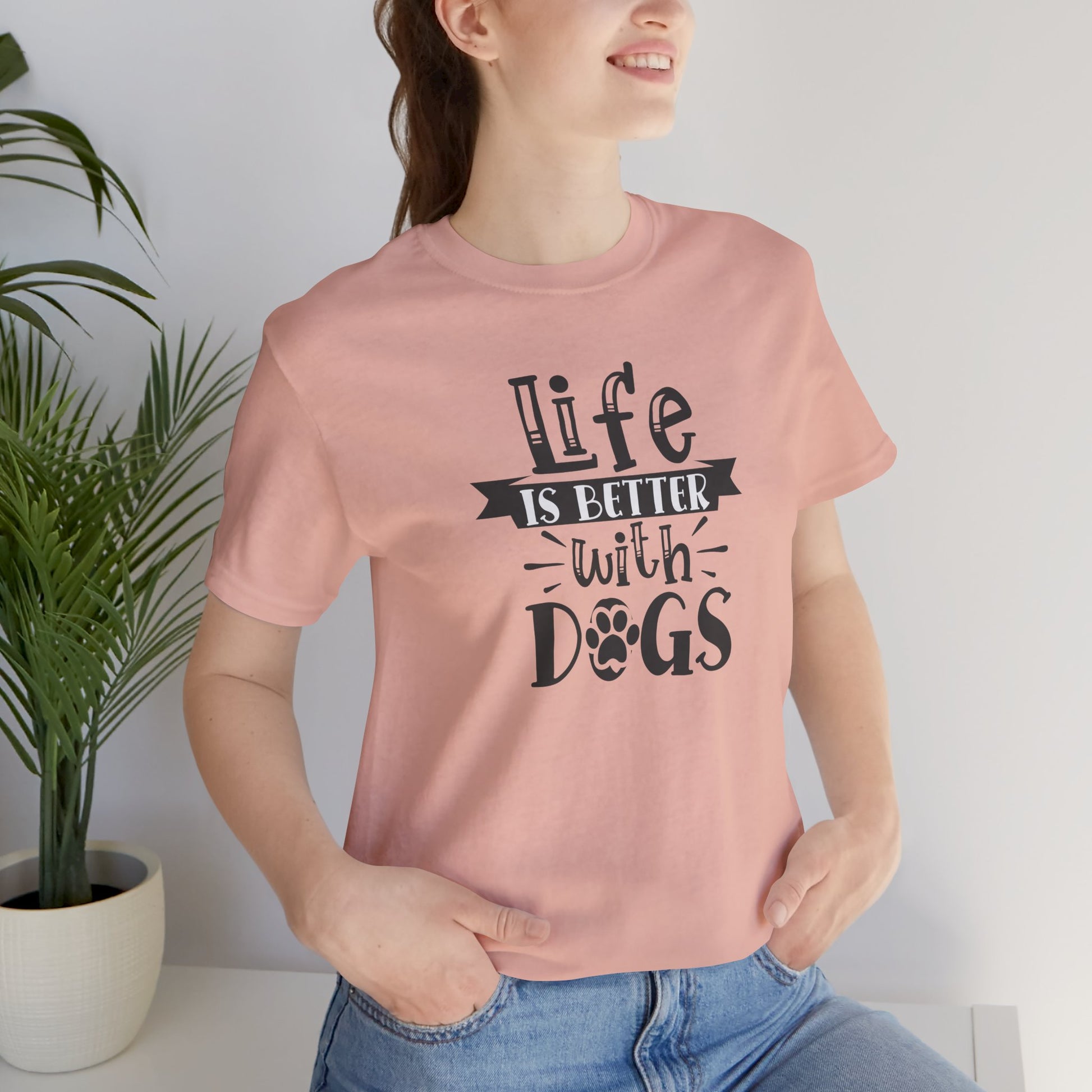Person wearing a pink t-shirt with 'Life is better with dogs' text, standing next to a plant.