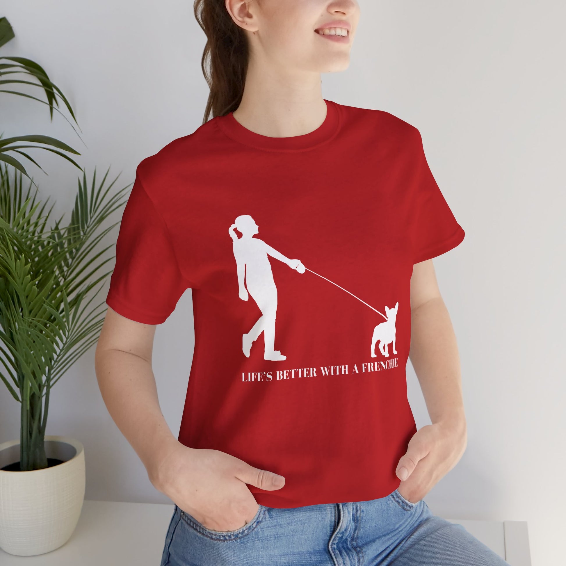 Person wearing a red t-shirt with a graphic and text, standing indoors next to a plant.